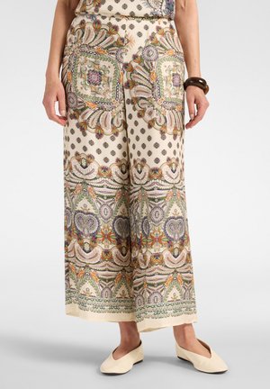 Femme portant un pantalon à jambes larges avec des motifs paisley et floraux multicolores complexes sur fond crème, associé à des ballerines crème.