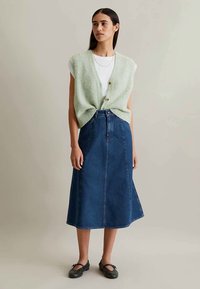 Vrouw in een lichtgroene mouwloze cardigan over een witte top, blauwe denim midi rok en zwarte Mary Jane schoenen, staand tegen een neutrale achtergrond.