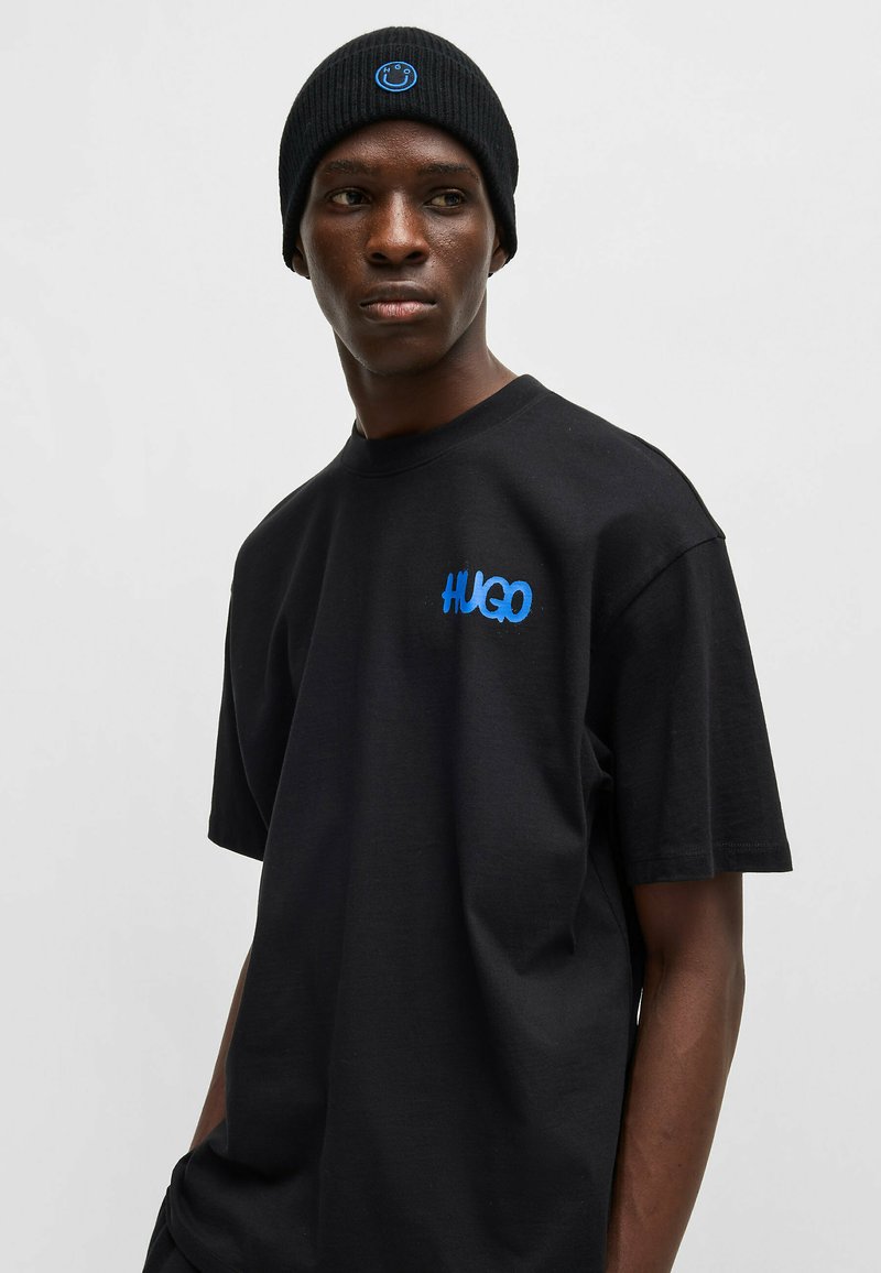 Zwarte katoenen T-shirt met ronde hals en korte mouwen, voorzien van het blauwe "HUGO" logo op de borst. Gemaakt met een zwarte geribde beanie.