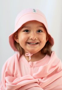 Rosa bucket-hatt med platt skärm och hakrem, matchande rosa zip-up plagg med slät textur och mjukt tyg. Barn som ler.