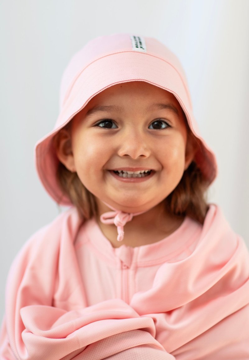 Rosa bucket-hatt med platt skärm och hakrem, matchande rosa zip-up plagg med slät textur och mjukt tyg. Barn som ler.