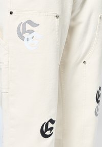 Pantalones cargo crema fabricados con un tejido grueso, que presentan logotipos bordados en negro y gris, detalles metálicos y bolsillos funcionales.