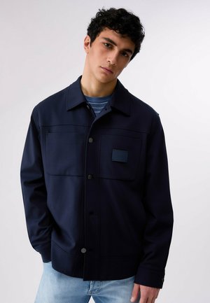 Ung mand med kort mørkt krøllet hår iført en mørkeblå jakke med knapper over en blåstribet skjorte og lyseblå jeans mod en ensfarvet baggrund.
