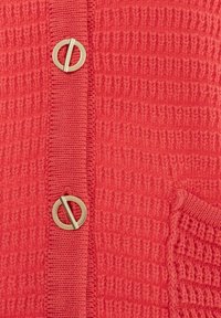 Cardigan texturé rouge avec un bouton circulaire doré proéminent, des bords côtelés et une poche en tricot sur le côté.