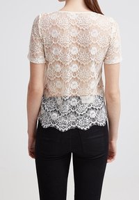 Top de encaje con mangas cortas, con un dobladillo ondulado, que pasa de blanco en la parte superior a negro en la parte inferior, mostrando patrones florales.