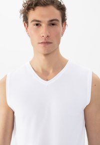 Jonge man met kort krullend haar in een witte mouwloze V-hals shirt, recht vooruit kijkend tegen een plain witte achtergrond.