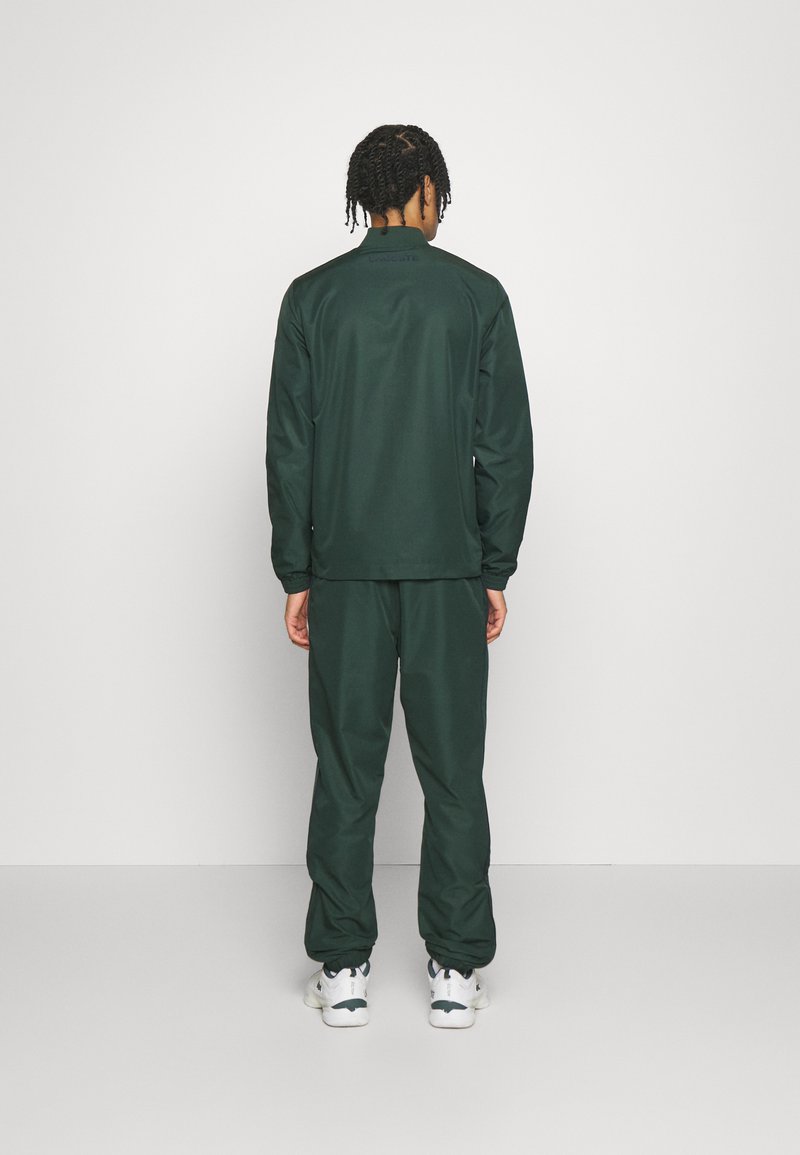 Hombre de pie de espaldas, vistiendo un chándal verde oscuro y zapatillas blancas, contra un fondo gris claro liso.