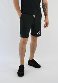 Pantalones cortos deportivos negros de mezcla de algodón, con cintura elástica y cordón, y un logo blanco. Usados con zapatillas negras y una camiseta negra.