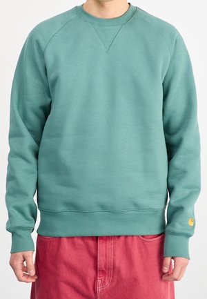 Mann trägt türkisfarbenen Sweatshirt mit gerippten Bündchen und Saum, kombiniert mit roten Hosen, steht vor einem schlichten hellen Hintergrund.