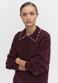 Bordowy kardigan o teksturowanym splocie, z kołnierzem ozdobionym dekoracyjnymi perłami i zapięciem na guziki, długie rękawy z ściągaczami.