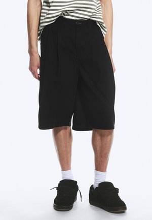 Partea inferioară a unei persoane care poartă pantaloni scurți largi negri, șosete albe, adidași negri și o cămașă cu dungi băgată în pantaloni.