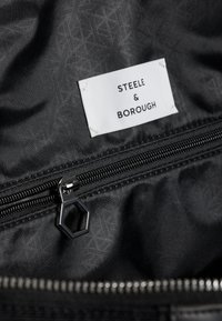 Černý interiér tašky s etiketou "STEEL & BOROUGH," obsahující zipovou kapsu s šestibokým zipem a vzorovanou látkou.