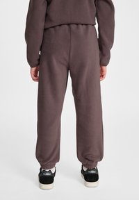 Fleece Jogginghose in Dunkelbraun, mit elastischen Bündchen und einem elastischen Bund. Getragen mit schwarzen Sneakers, die von weißen Sohlen akzentuiert sind.