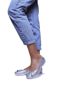 Zapatos planos plateados de tejido con punta redondeada, que presentan un pequeño lazo como acento. Llevados con unos pantalones vaqueros de mezclilla clara y remangados.