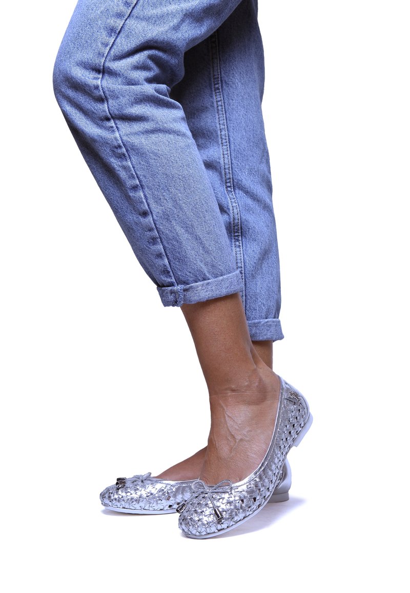 Zapatos planos plateados de tejido con punta redondeada, que presentan un pequeño lazo como acento. Llevados con unos pantalones vaqueros de mezclilla clara y remangados.