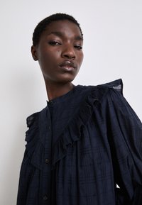 Navyblaue Bluse mit Knöpfen aus strukturierter Stoffqualität, mit Rüschendetails an den Schultern und einem Kragen. Besitzt ein kariertes Muster und eine normale Passform.