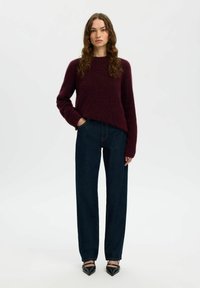 Donna con capelli ricci e lunghi che indossa un maglione bordeaux, jeans dritti blu scuro e décolleté neri con punta a stiletto, in piedi davanti a uno sfondo bianco.
