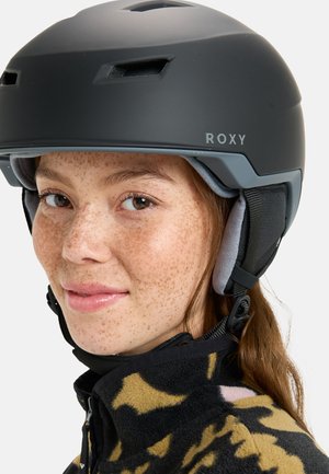 Casque de ski noir avec des fentes de ventilation, marquage Roxy et coussinets d'oreilles doux. Le modèle porte une veste en polaire à motifs en dessous.