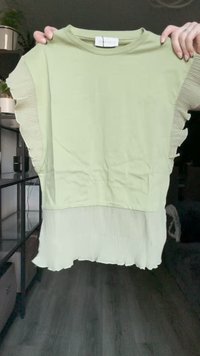T-shirt verde claro de manga curta com mangas em camadas pregueadas com bainha canelada, segurada por duas mãos em ambiente interno.