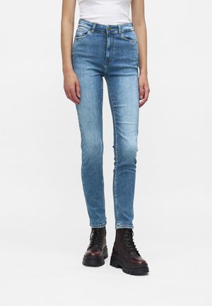 Personne portant un jean skinny taille haute bleu clair et des bottes de combat marron foncé à lacets, debout sur un fond blanc uni.