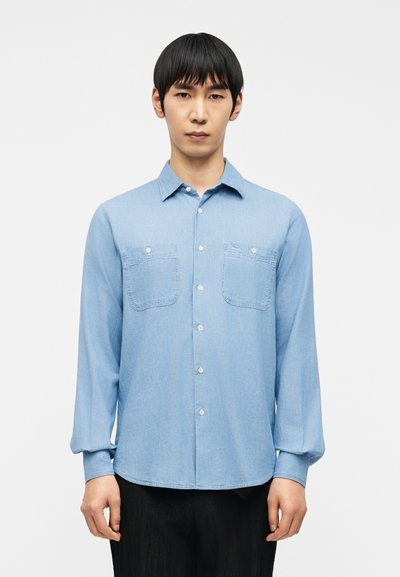 Jeune homme aux cheveux noirs raides portant une chemise bleu clair à boutons avec deux poches poitrine et un pantalon noir, sur un fond uni.