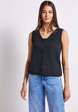 Schwarze, texturierte, ärmellose Weste mit V-Ausschnitt und Knopfverschluss, kombiniert mit hellblauen, lässig geschnittenen Jeans. Schlichtes Design und lässiger Look.