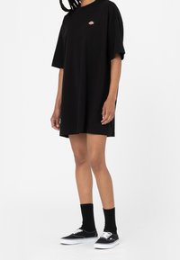 Osoba ubrana w luźną czarną sukienkę T-shirt, czarne skarpetki i czarne płócienne trampki z białymi podeszwami i sznurówkami, stojąca na białym tle.