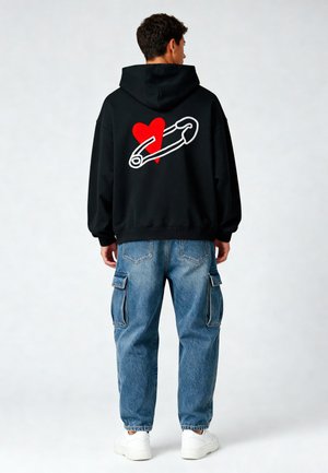 Felpa nera con grafica di un cuore rosso e un fermaglio di sicurezza bianco sul retro, abbinata a jeans cargo azzurri e sneakers bianche.