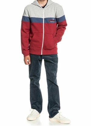 Man in een zip-up hoodie met grijze, marineblauwe en bordeauxrode kleurblokken, donkere jeans en witte sneakers, staande met één hand in zijn zak.