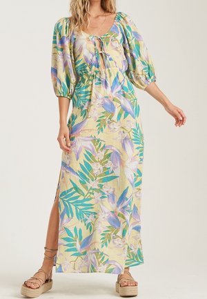 Vrouw die een lange bloemige jurk draagt met pofmouwen, details met strik aan de voorkant, een zijsplit en platte sandalen met enkelbandjes.