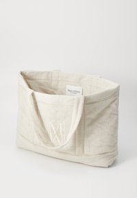Sac fourre-tout en toile beige avec une finition texturée, doté de deux poignées robustes et d'un détail en relief du logo 'M' à l'avant. Étiquette intérieure visible.