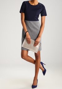 Kobieta w granatowej bluzce i wzorzystej spódnicy trzyma beżową kopertówkę, ma na sobie granatowe szpilki, stoi z jedną nogą skrzyżowaną.