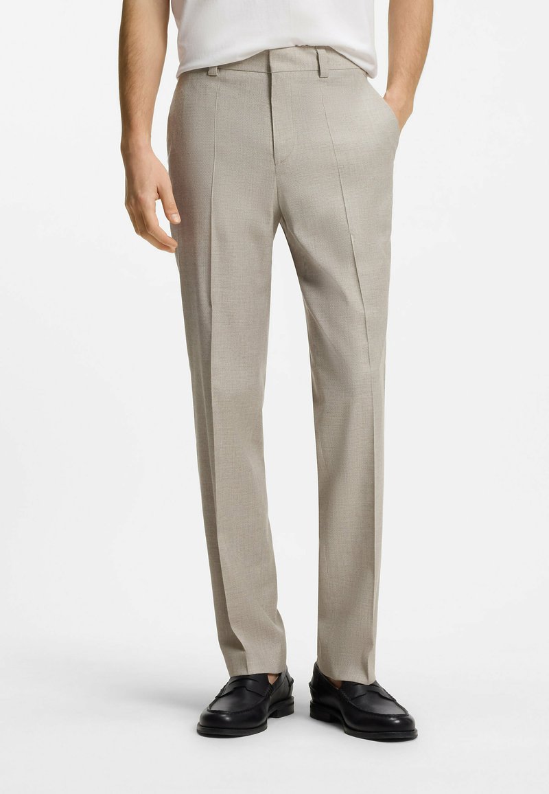 Homme portant un pantalon beige sur mesure et des mocassins en cuir noir, debout, les mains détendues, sur un fond blanc uni.