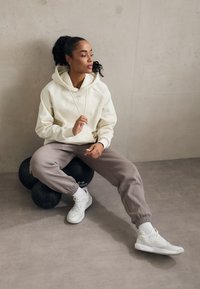 Felpa crema con cordini, pantaloni della tuta grigi con polsini elasticizzati e scarpe da ginnastica bianche. Seduti su palle da esercizio nere contro un muro neutro.