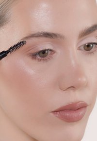 Schwarzer Augenbrauengel-Applikator, der durch hellbraune Augenbrauen gestrichen wird. Sanfte, natürliche Hautfarbe mit einem weichen, glänzenden Nude-Lippenstift. Subtile Augen-Make-up.