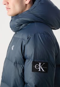 Marineblaues Puffer-Jacke mit glänzender Textur, ausgestattet mit einer großen Kapuze und einem Logo-Patch am Ärmel. Steppdesign mit sichtbaren Nähten.