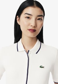 Vit polotröja med marinblå krage och knappslå. Har en grön broderad krokodil-logotyp på vänster bröst. Strukturerad tyg.