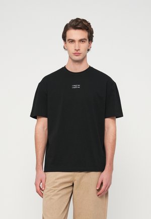 Junger Mann, der nach vorne schaut und ein schwarzes T-Shirt mit dem Schriftzug "LIBERTINE LIBERTINE" sowie beige Hosen trägt, vor einem schlichten Hintergrund.