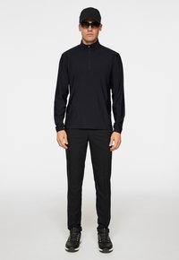 Maglia nera a maniche lunghe con zip e colletto alto, abbinata a pantaloni neri aderenti e scarpe da ginnastica nere. Include un cappellino nero e occhiali da sole.