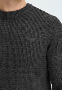 Mørkegrå strikket sweater med et struktureret, ribbet mønster, der har en rund halsudskæring og et lille logo mærke på fronten.