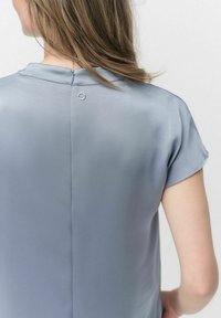 Kortærmet, lysegrå bluse med en draperingsstruktur, der har en nøglehulslukning bagpå og subtil syning langs udskæringen.