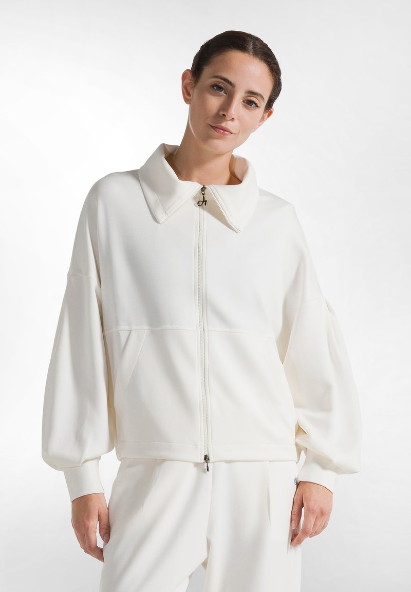 Chaqueta blanca de tejido suave con cremallera y gran cuello, hombros caídos y bolsillos laterales. Presenta un sutil brillo y una silueta relajada.