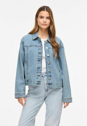 Kvinde iført lyseblå cowboyjakke over hvid top og lyseblå jeans, stående mod en ensfarvet hvid baggrund.