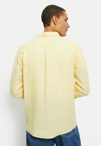 Helles gelbes Langarmhemd aus Leinenstoff mit Rückenpasse, Button-Down-Kragen und abgerundetem Saum. Getragen mit blauen Denim-Hosen.