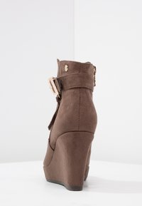 Scarpa con zeppa in suede marrone con punta rotonda, cinturino alla caviglia e un accento in oro sulla fibbia. Texture liscia e tacco leggermente elevato.
