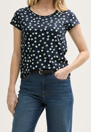 Granatowy t-shirt z krótkim rękawem w zielony wzór w kropki; okrągły dekolt; noszony z niebieskimi dżinsami i czarnym paskiem.