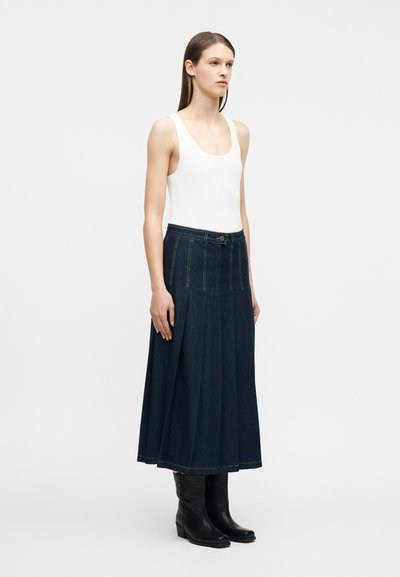 Γυναίκα που στέκεται με λευκό αμάνικο τοπ, σκουρό μπλε πλισέ τζιν midi φούστα και μαύρες μπότες μέχρι τη μέση της γάμπας, μπροστά σε απλό φόντο.