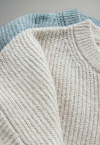Zwei überlappende gestrickte Pullover, einer in Creme mit vertikalen Rippen und einer in Hellblau mit strukturierter Oberfläche, beide mit Rundhalsausschnitt.