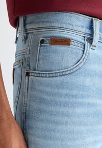 Κοντινή λήψη ανοιχτό μπλε τζιν παντελονιού που δείχνει την μπροστινή τσέπη με καφέ ετικέτα λογότυπου Wrangler και μέρος από το χέρι ενός ατόμου με κόκκινη μπλούζα.