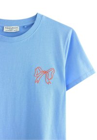 Vaaleansininen lyhythihainen puuvillapaita, jossa on pyöreä pääntie ja pieni korallinvärinen brodeerattu solmu vasemmassa rintataskussa.